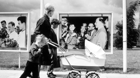 Ein Mutter geht Anfang der 60er-Jahre mit ihren Kindern an Fotowänden mit SED-Propaganda, unter anderem mit Walter Ulbricht im Bild, vorbei. © picture-alliance / akg-images / Gardi Foto: Gardi